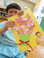 色画用紙に、色をつけたシールで作った花束を描いた作品を持つ幼児